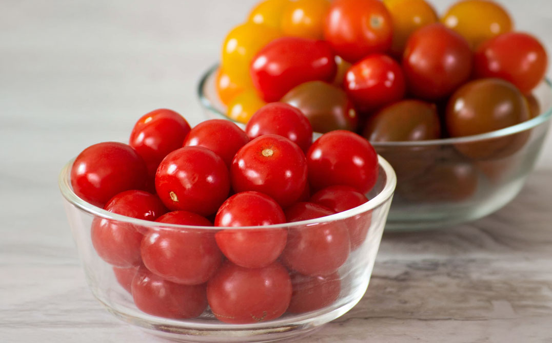 Two glass bowls filled with cherry tomatoes. The bowl in front contains red tomatoes, while the bowl in the background has a mix of yellow, red, and brown tomatoes. Both bowls are placed on a light-colored surface. GREENHOUSE Goodness