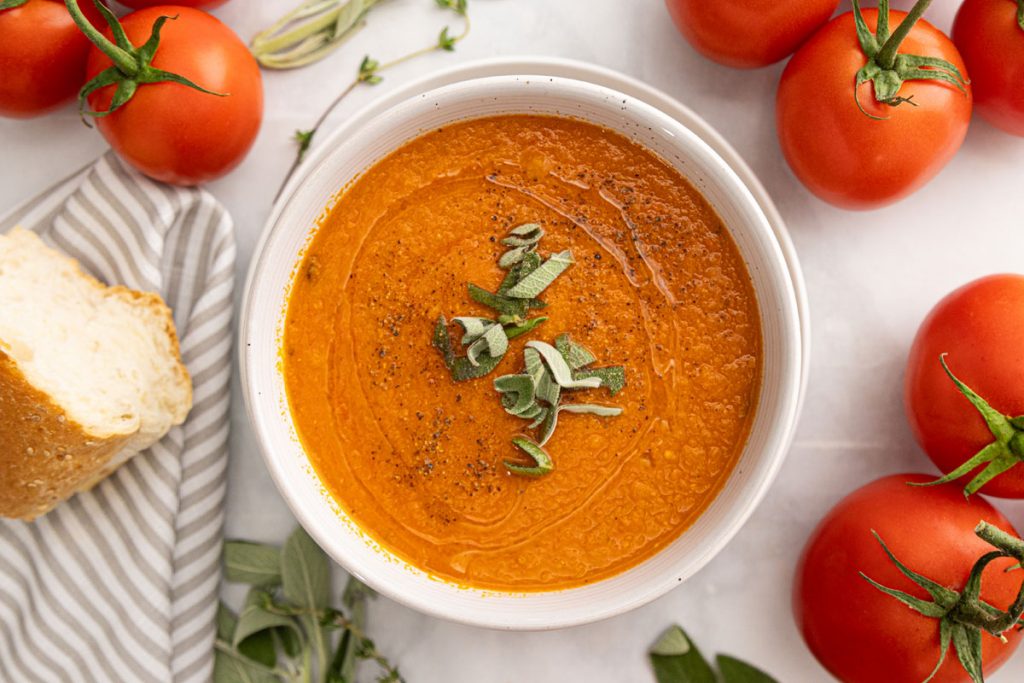 A white bowl of tomato soup garnished with fresh herbs sits on a light surface, surrounded by vine tomatoes, a slice of bread on a striped napkin, and sprigs of fresh herbs. GREENHOUSE Goodness