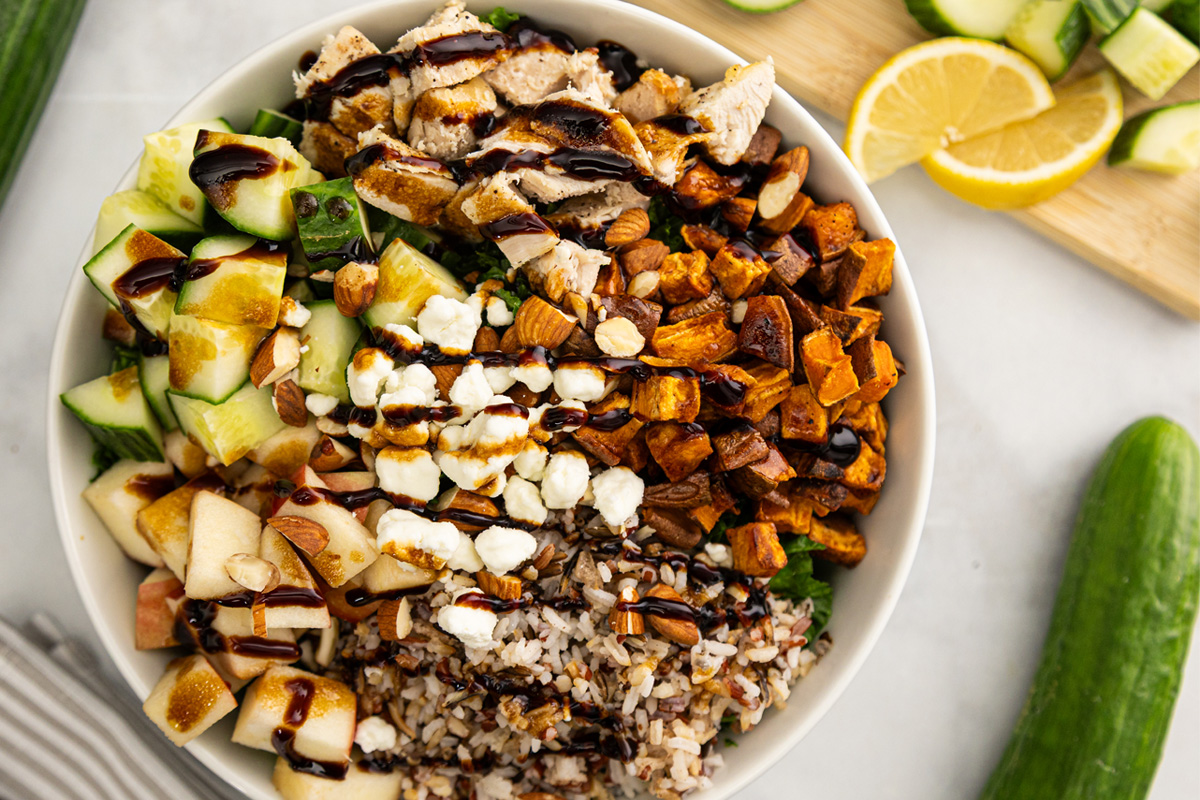 A bowl filled with chopped cucumbers, grilled chicken, sweet potatoes, apples, wild rice, almonds, goat cheese, and drizzled with dark balsamic glaze; lemon slices and cucumber are on the side. GREENHOUSE Goodness