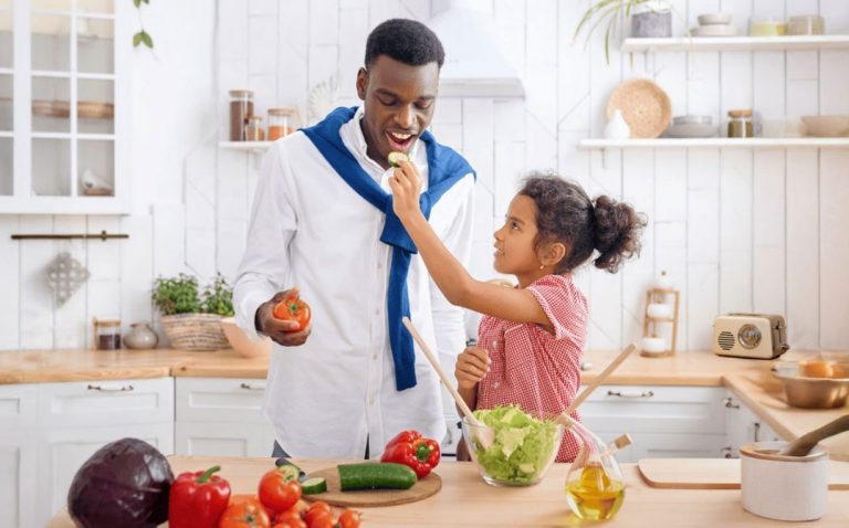 A young girl feeds a man a slice of cucumber in a bright kitchen. They smile while preparing a fresh salad with various vegetables on the counter. GREENHOUSE Goodness