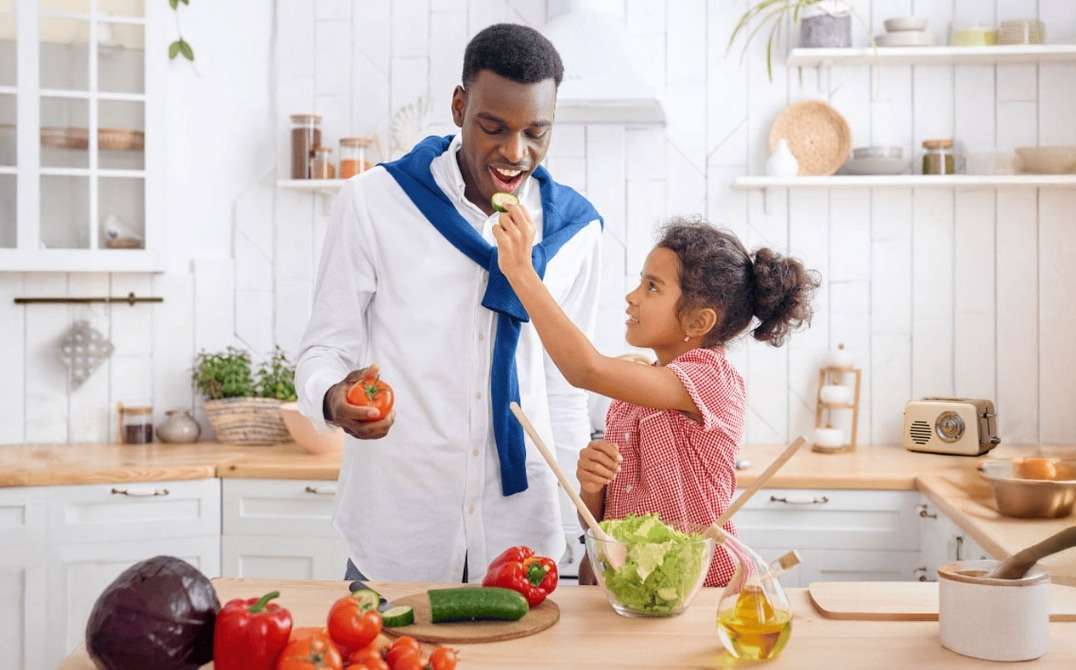 A young girl feeds a man a slice of cucumber in a bright kitchen. They smile while preparing a fresh salad with various vegetables on the counter. GREENHOUSE Goodness