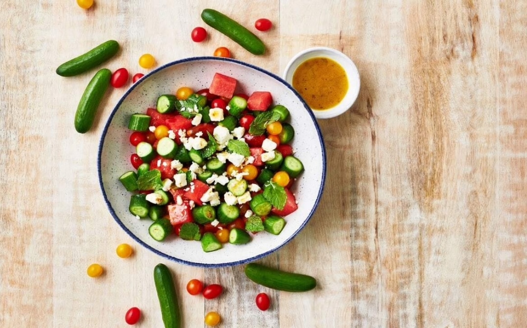 A bowl of fresh salad featuring greenhouse cucumber recipes with watermelon, cherry tomatoes, feta cheese, and mint leaves. Small cucumbers and tomatoes are scattered around, with a small bowl of dressing on the side. GREENHOUSE Goodness