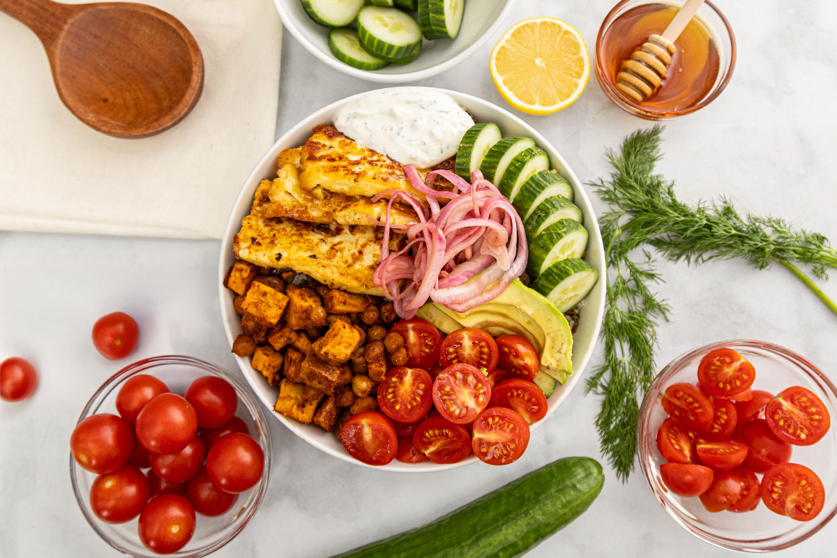A bowl filled with grilled halloumi, roasted sweet potatoes, chickpeas, sliced cucumber, avocado, cherry tomatoes, pickled onions, and tzatziki, surrounded by bowls of vegetables, honey, lemon, and fresh herbs on a white table. GREENHOUSE Goodness