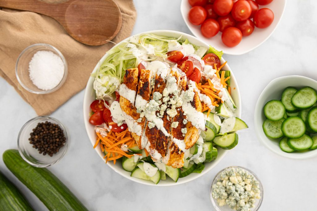 A bowl of salad with grilled chicken, blue cheese crumbles, cherry tomatoes, cucumbers, shredded carrots, and lettuce, surrounded by small bowls of salt, pepper, tomatoes, blue cheese, and sliced cucumbers on a white surface. GREENHOUSE Goodness