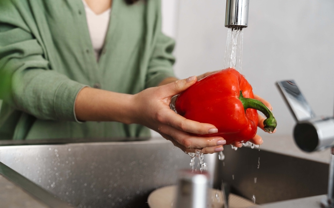A person in a green shirt washes a red bell pepper under running water in a kitchen sink. GREENHOUSE Goodness