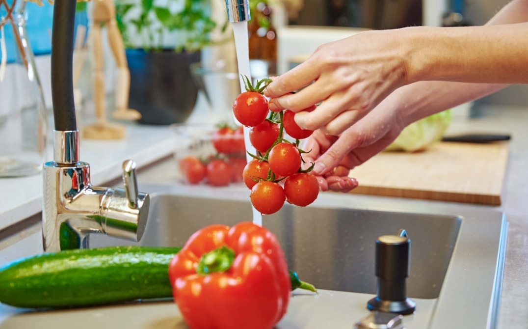 A person washes a bunch of cherry tomatoes under running water in a kitchen sink. A cucumber and a red bell pepper are on the counter nearby, with other vegetables and kitchen items visible in the background. GREENHOUSE Goodness