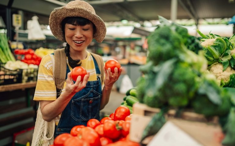 A woman wearing a straw hat and overalls smiles while holding two tomatoes at a market stall filled with fresh vegetables like tomatoes, broccoli, and cauliflower. GREENHOUSE Goodness
