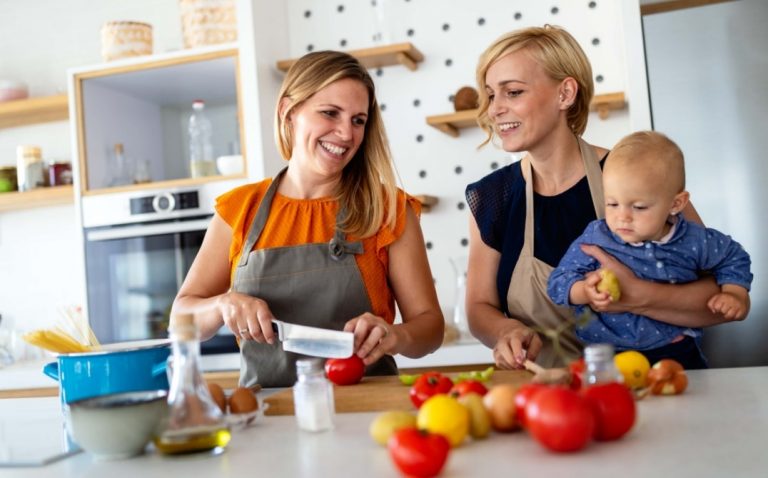 Two women in aprons smile while preparing food in a kitchen. One chops vegetables, and the other holds a baby who is holding a yellow fruit. Fresh produce and cooking ingredients are spread on the counter. GREENHOUSE Goodness