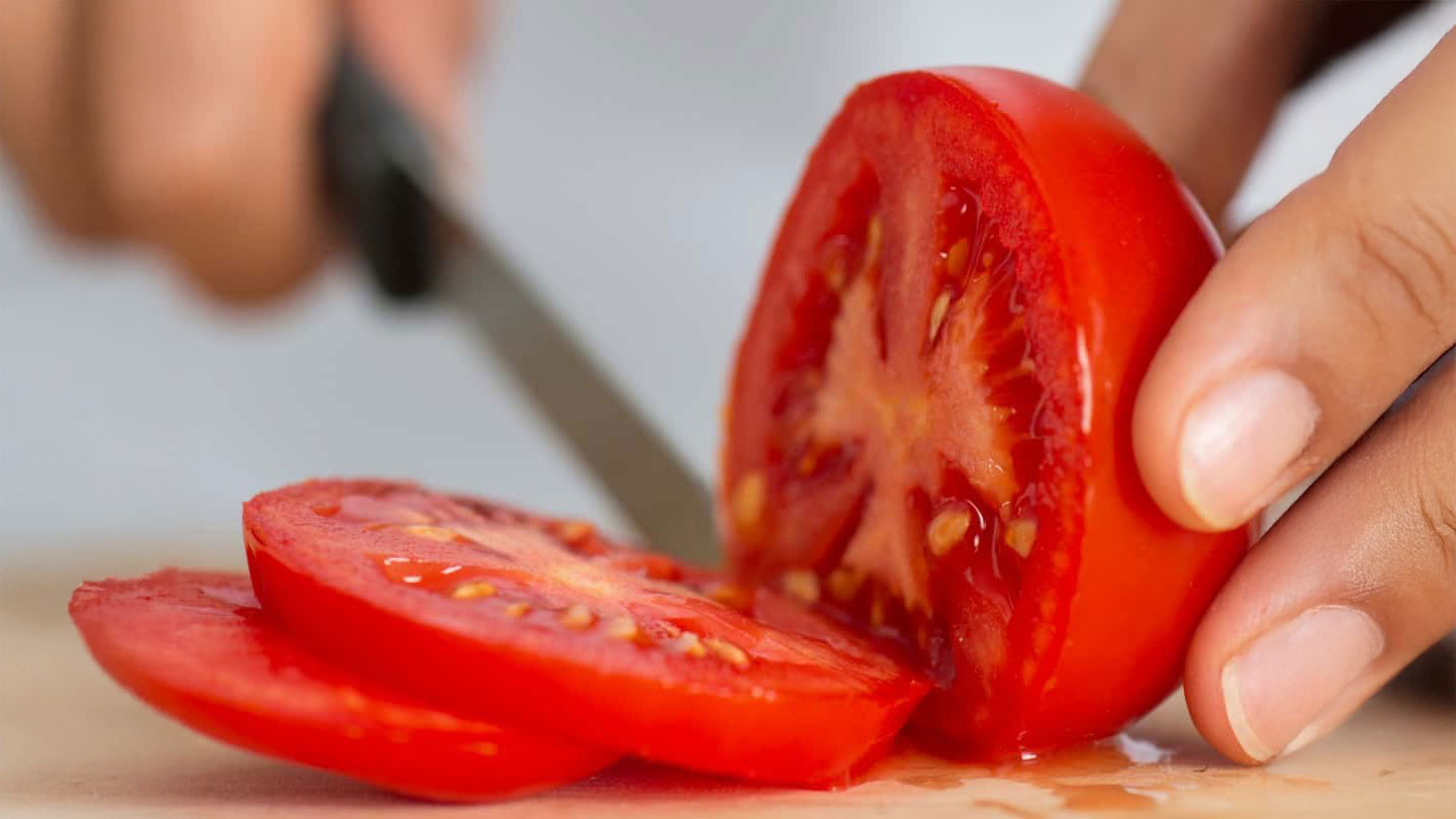 Close-up of a hand slicing a ripe red tomato with a knife on a cutting board; several tomato slices are visible in the foreground. GREENHOUSE Goodness