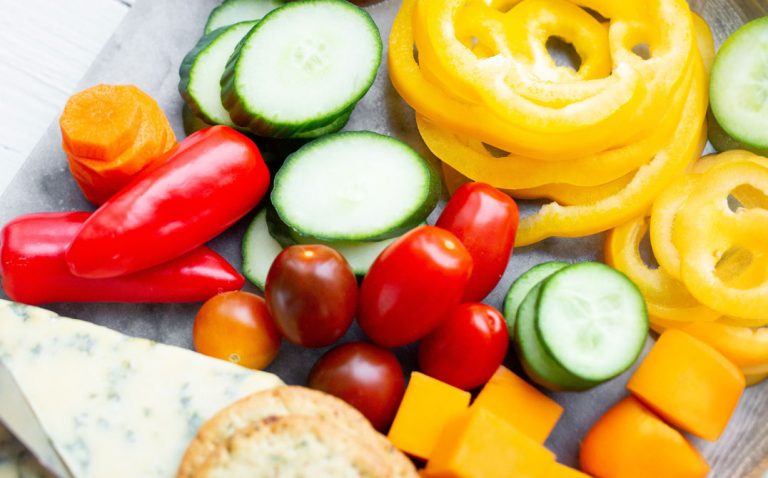 A colorful assortment of fresh vegetables, including sliced yellow bell pepper, cucumber, grape tomatoes, carrots, and red mini peppers, alongside cubes of cheese, crackers, and a wedge of blue cheese. GREENHOUSE Goodness