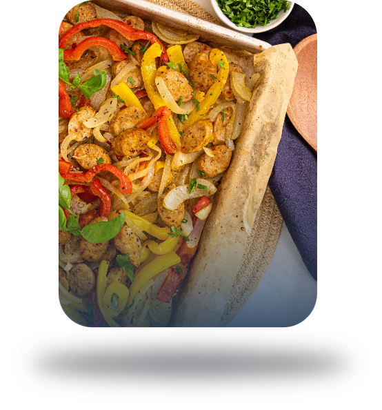 A sheet pan with cooked sausage slices, red and yellow bell peppers, and onions garnished with fresh herbs, served on parchment paper with a side dish of chopped greens in the corner. GREENHOUSE Goodness