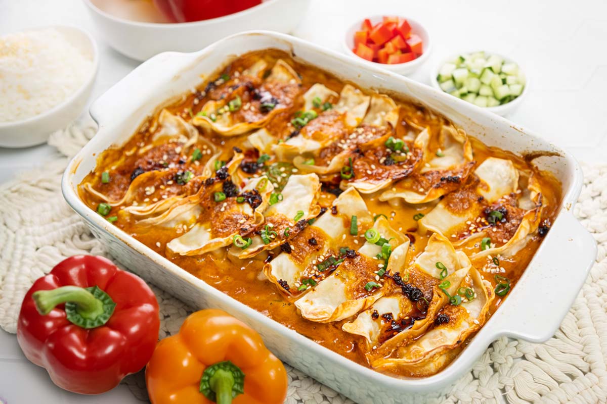 A baked casserole dish filled with dumplings in a rich orange sauce, garnished with chopped green onions. Surrounding the dish are fresh red and yellow bell peppers, and small bowls of diced vegetables. GREENHOUSE Goodness