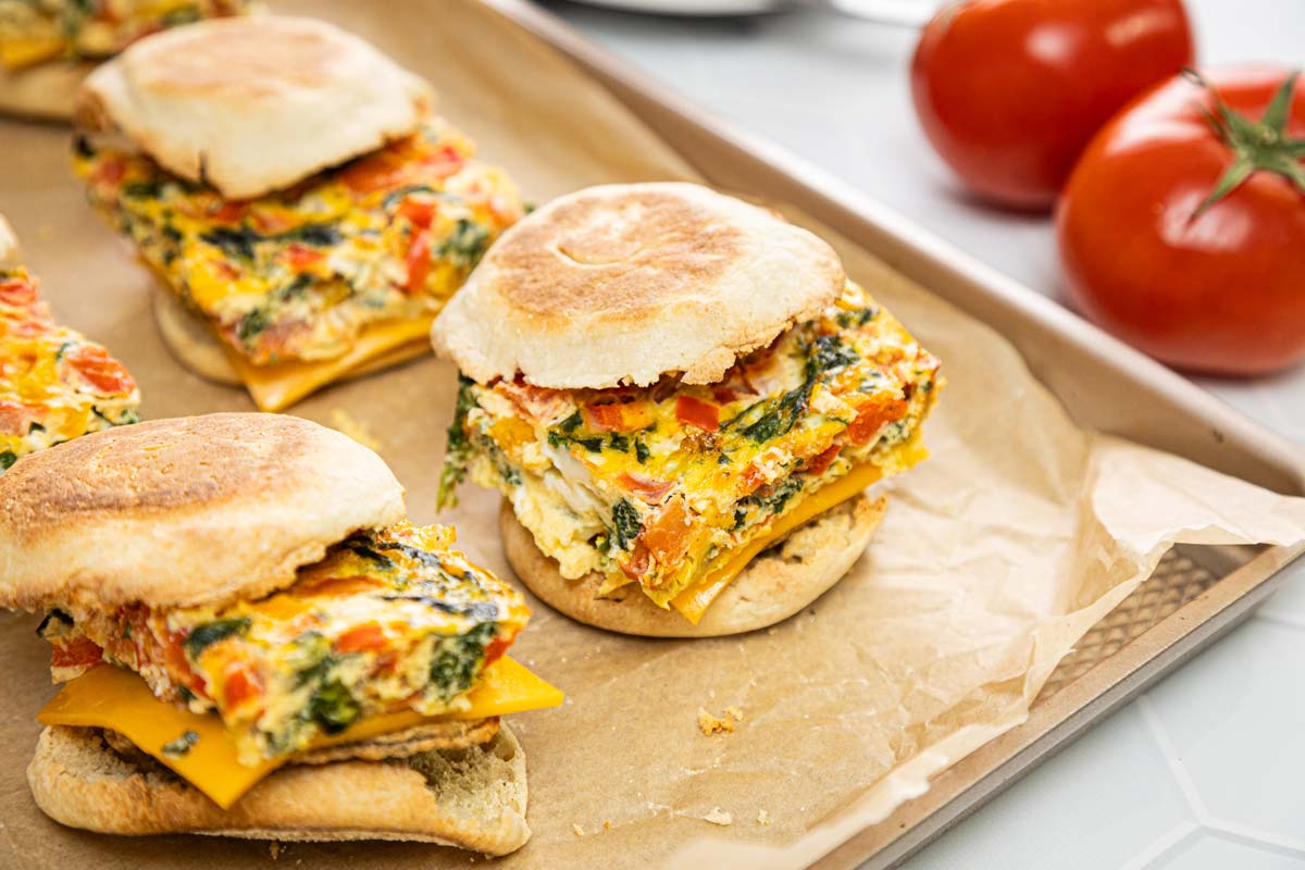 Breakfast sandwiches on English muffins filled with egg, spinach, tomato, and cheddar cheese, arranged on a parchment-lined baking sheet with whole tomatoes in the background. GREENHOUSE Goodness