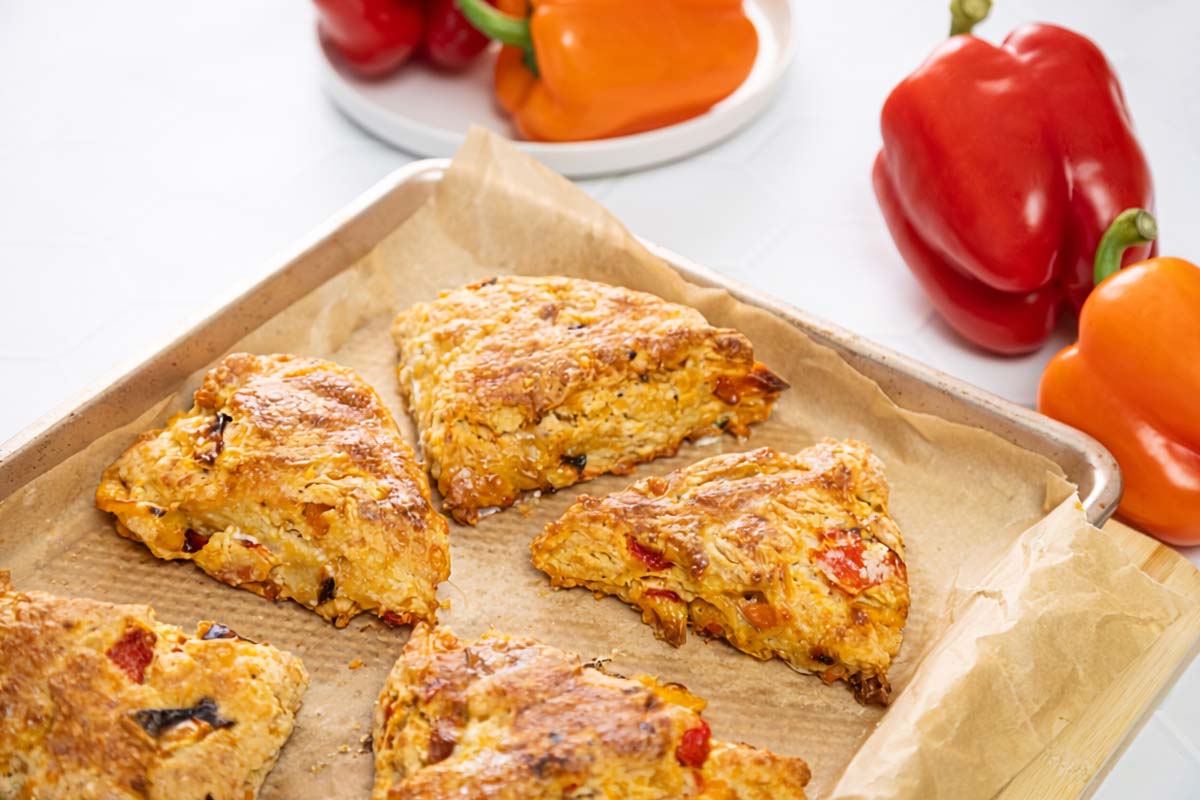 Golden-brown scones with pieces of red and orange bell pepper on a parchment-lined baking sheet. Fresh red and orange bell peppers are visible in the background on a white surface. GREENHOUSE Goodness