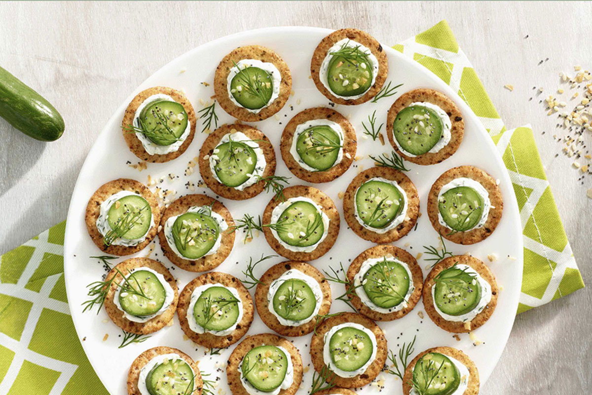 A white plate holds several round crackers topped with a layer of cream cheese, cucumber slices, dill, and seasoning. The plate sits on a green and white patterned napkin on a light-colored surface. GREENHOUSE Goodness